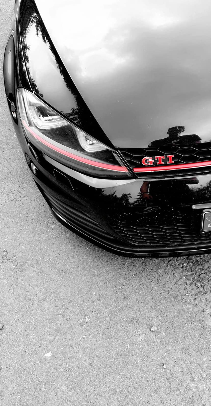 Volkswagen GTI front grille with a red stripe, silver badge, and honeycomb patterns reflected on the black hood.