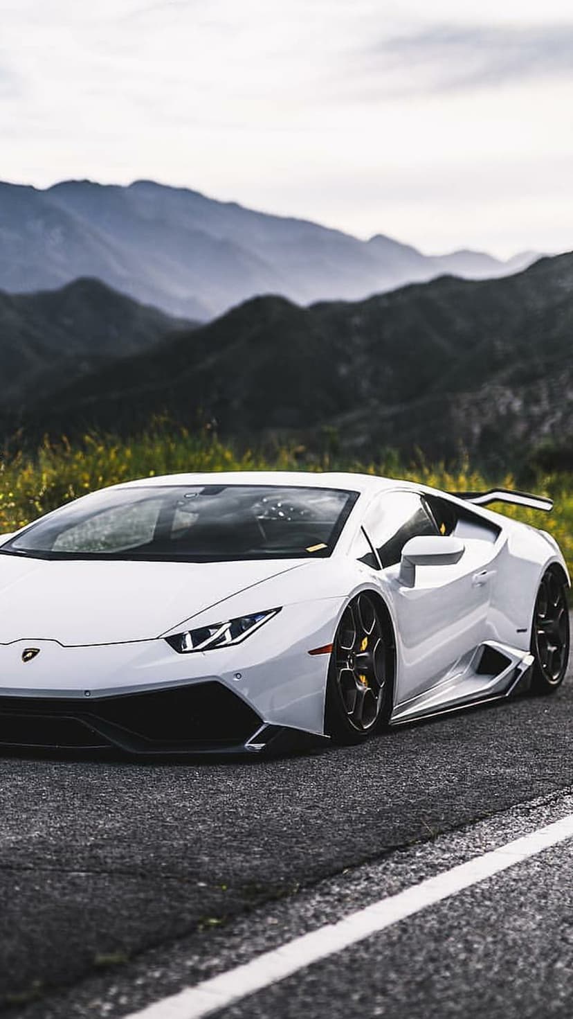 White Lamborghini supercar on a mountain road with green grass and towering misty peaks in the background.