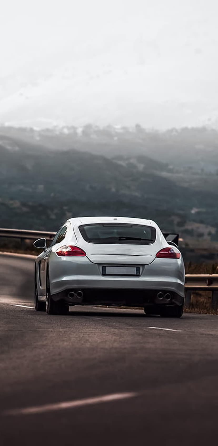 White Porsche Panamera rear profile driving on a winding mountain road through dense fog and evergreen trees.