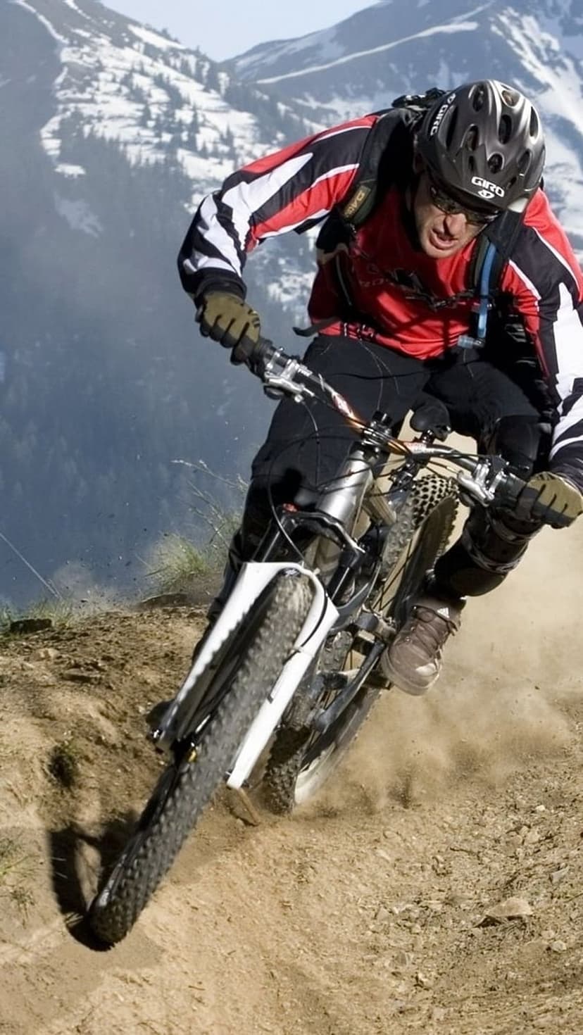 Mountain biker in full gear drifts on a dirt path with snow-capped mountains and pine forests in the distance.