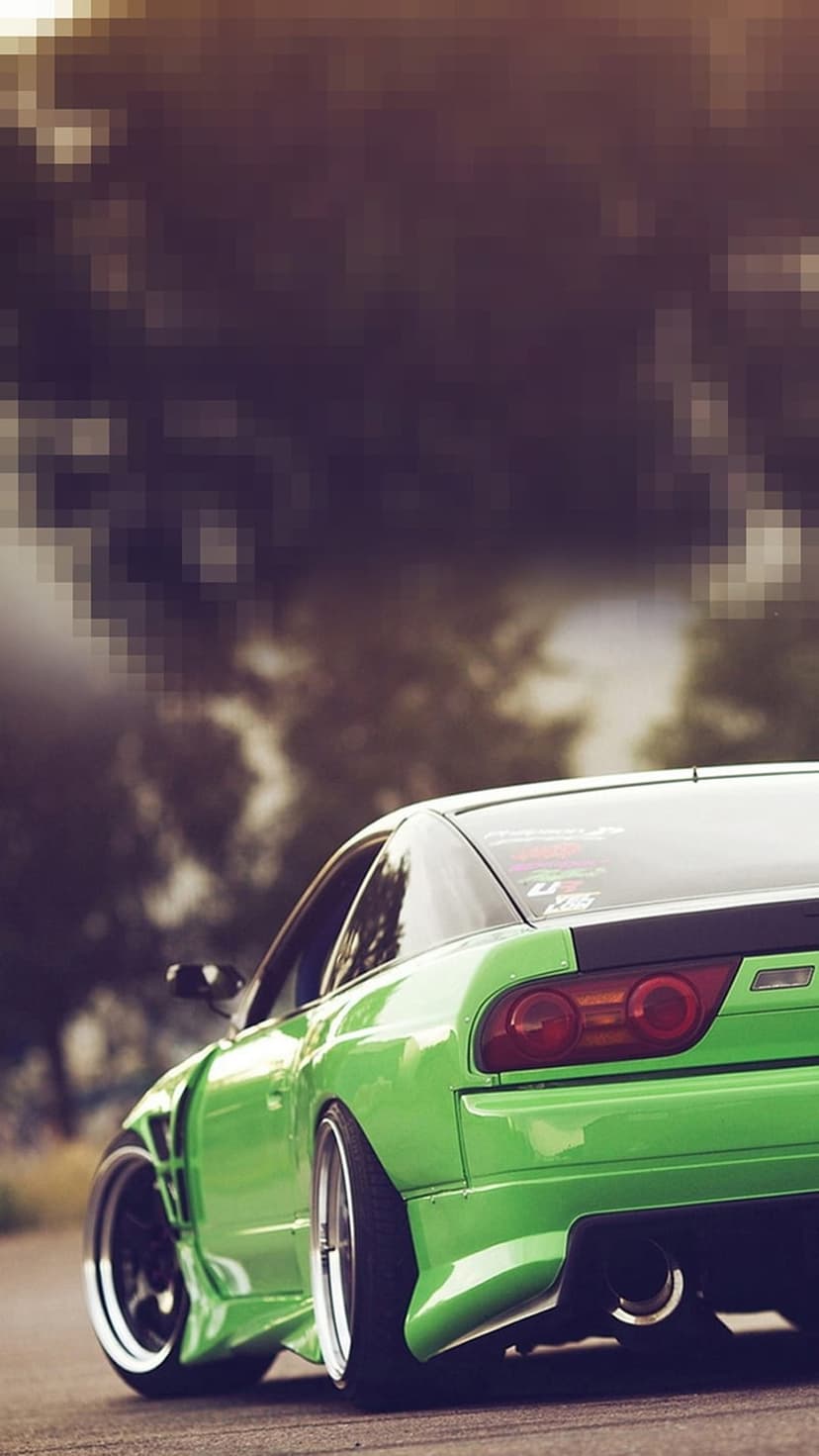 Green Nissan Silvia S13 with widebody fenders and chrome wheels parked on a road with a blurred forest background.