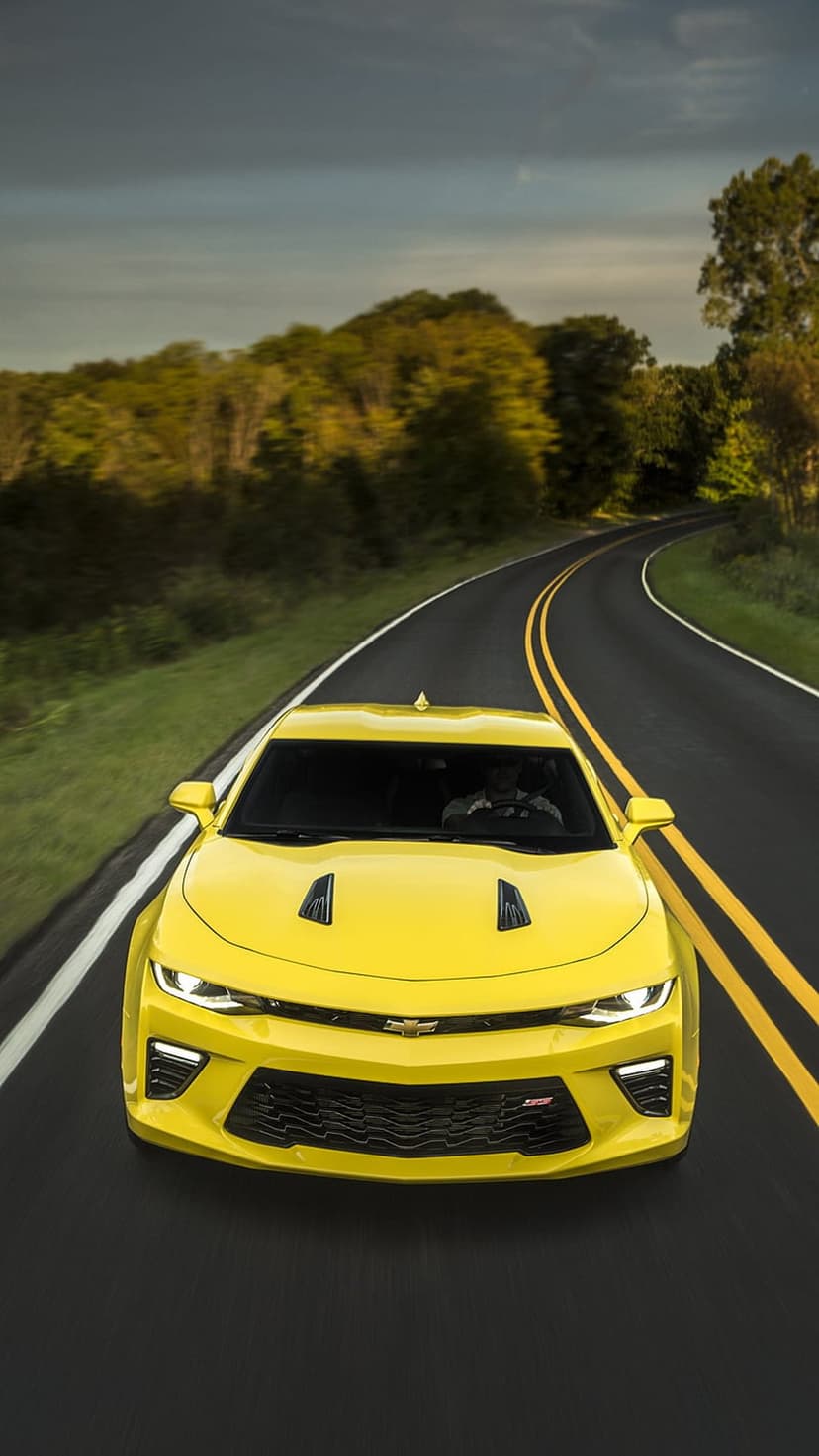Yellow Chevrolet Camaro travels down a winding road through green trees with glowing headlights and yellow lines.