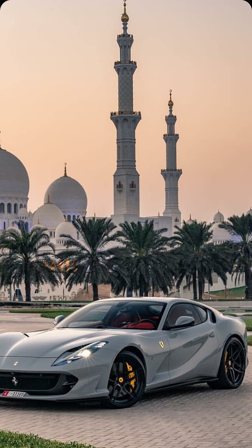 Silver Ferrari supercar with red interior parked before the illuminated white domes of a Dubai mosque at sunset.