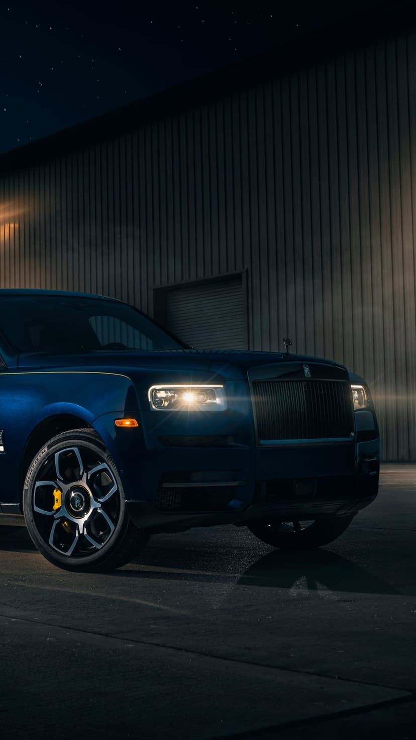 Deep blue luxury SUV with yellow accents and glowing headlights parked by an industrial wall at night.
