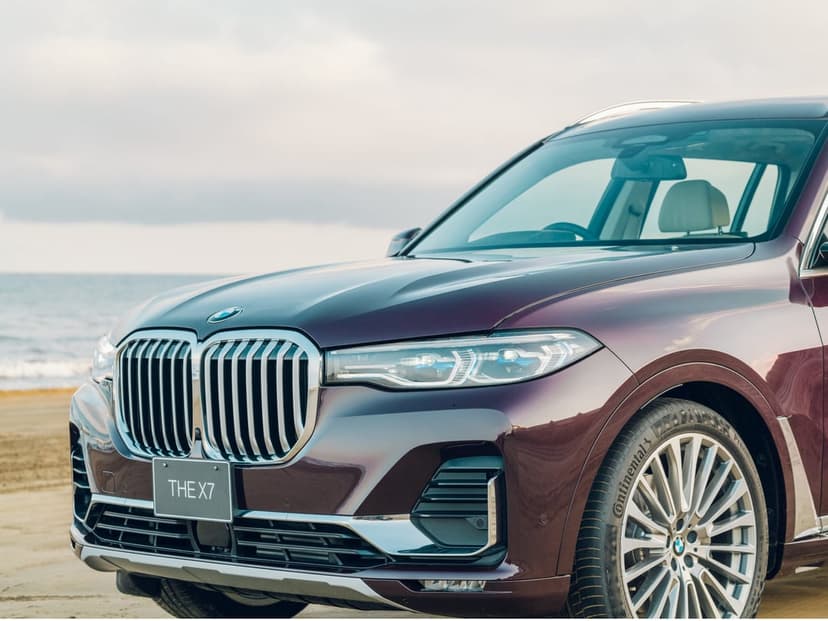 Luxury dark purple BMW X7 SUV parked on a sandy beach with the ocean in the background under a soft sky.