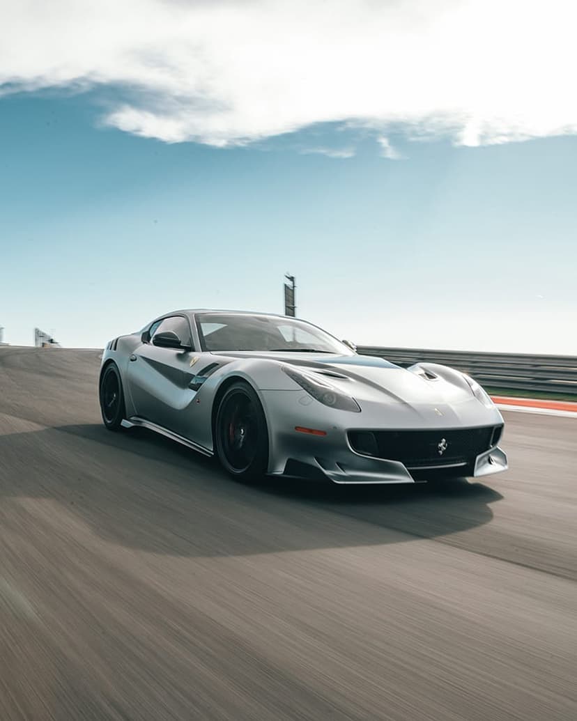 Silver Ferrari F12 TDF racing on a circuit with motion blur against a blue sky with clouds in a low-angle view.