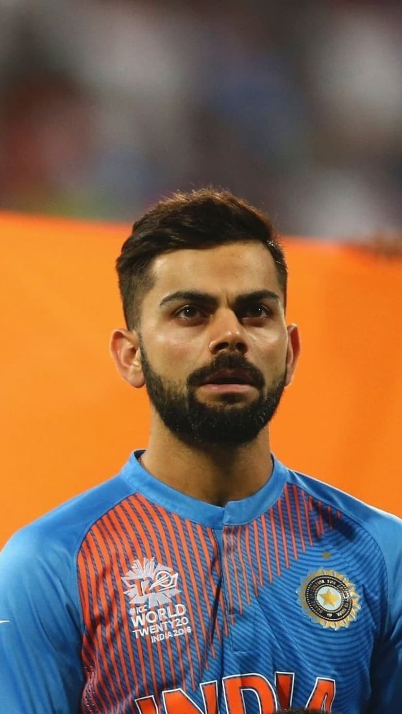 Virat Kohli in a blue India cricket jersey with orange collar details stares forward with a focused expression.