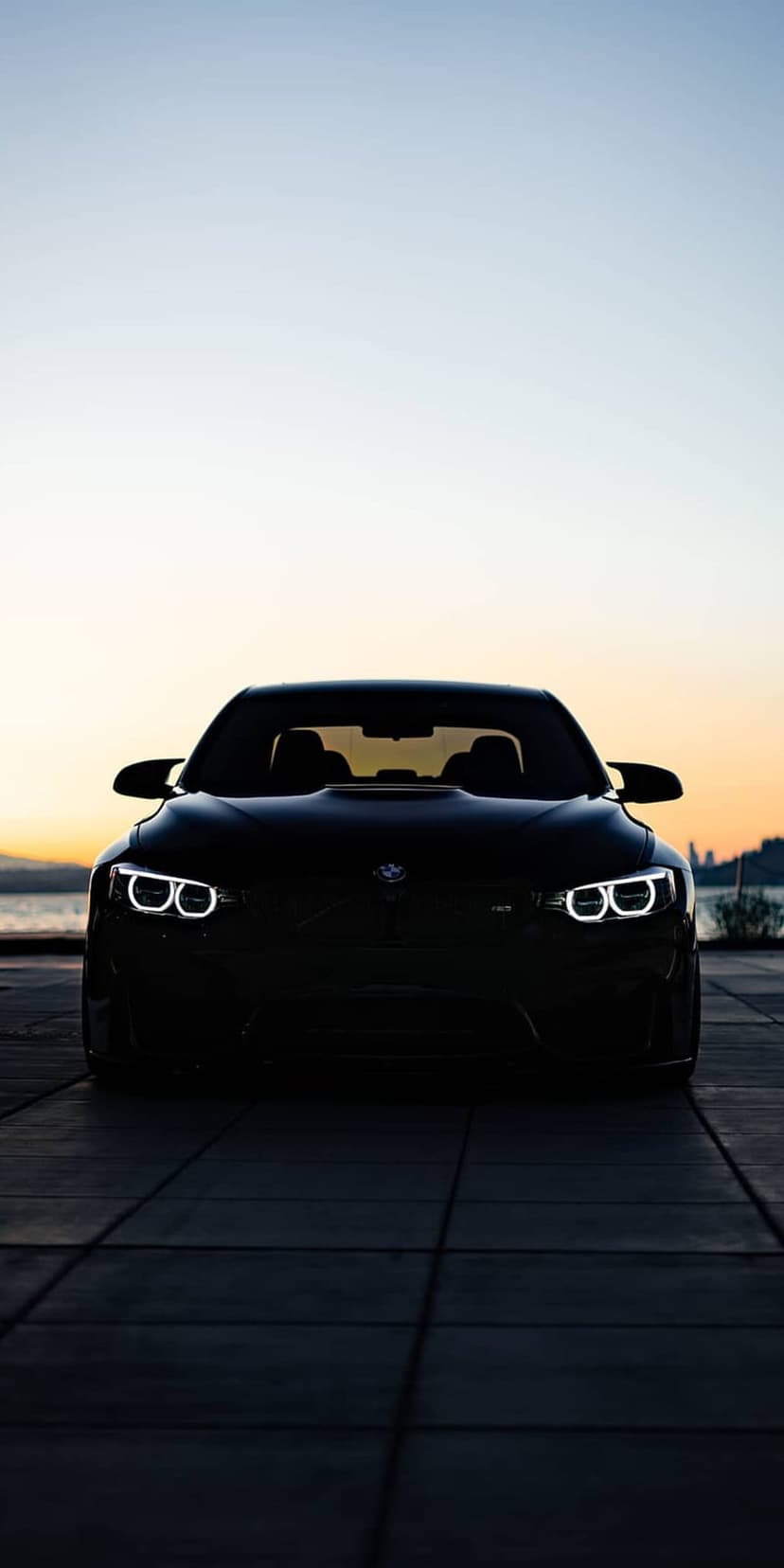 BMW M4 sports car with glowing LED headlights parked on a tiled surface during a vibrant city sunset.