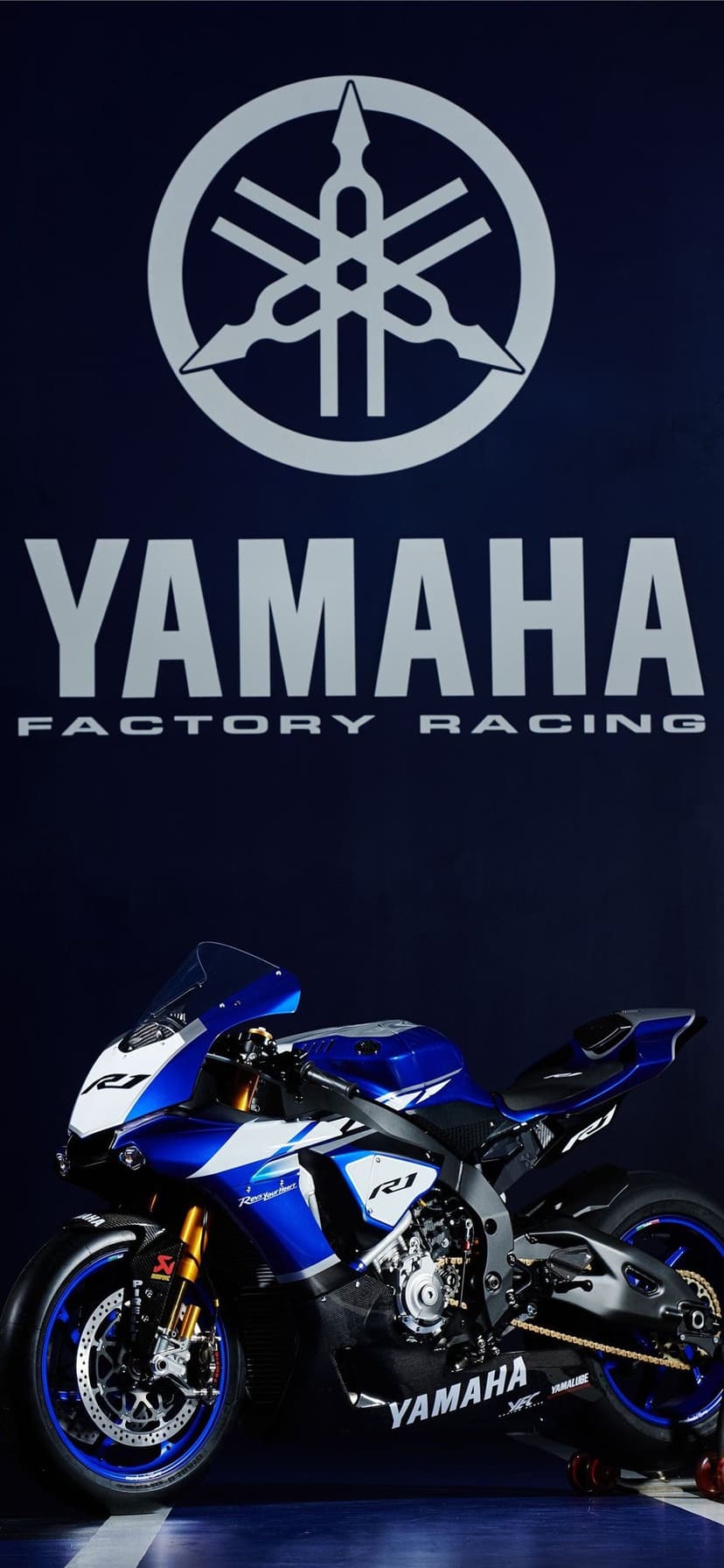 Yamaha YZF-R1 sportbike in blue racing livery parked beneath a large white tuning fork logo on a navy wall.