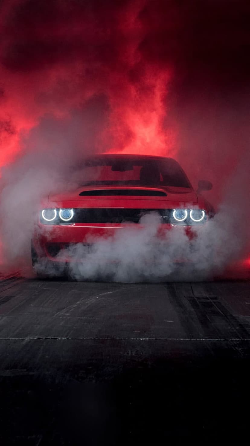 Red Dodge Challenger performing a burnout with thick white smoke and glowing headlights on dark asphalt.