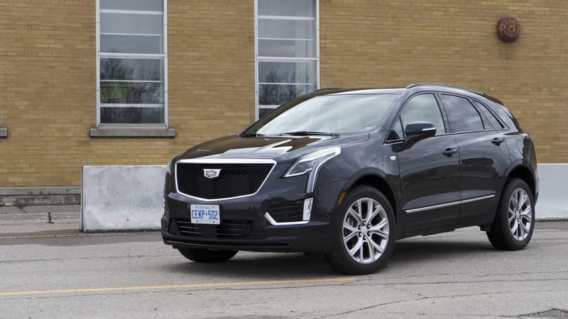 Dark Cadillac XT5 luxury SUV parked on grey asphalt in front of a textured tan brick wall with large windows.
