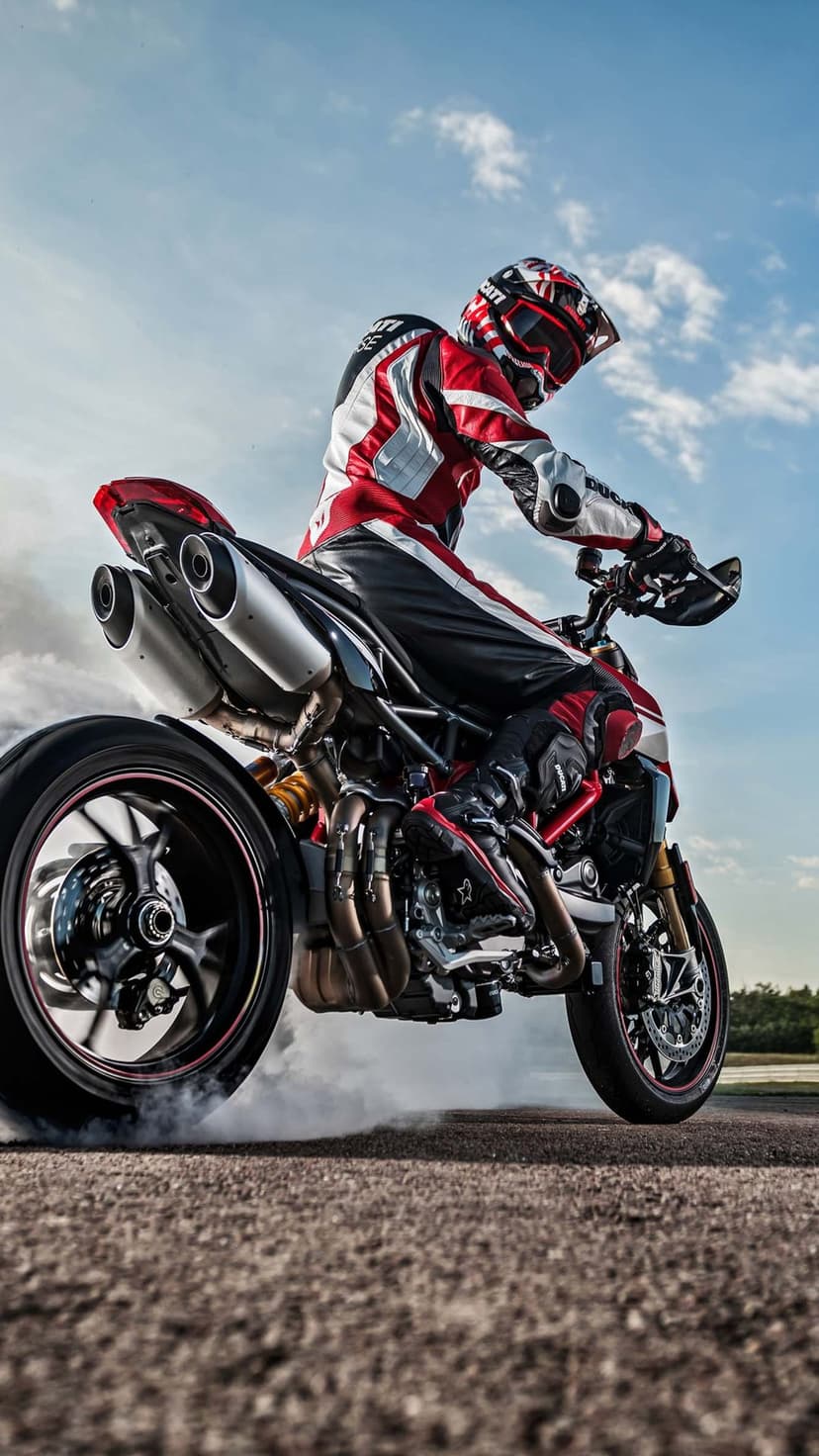 Motorcyclist in red racing gear performs a tire burnout on a Ducati superbike against a cloudy sky backdrop.