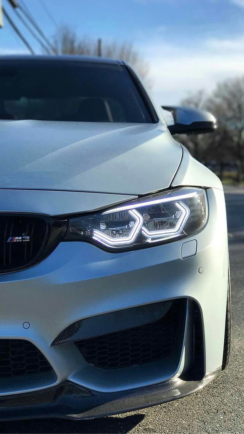 White BMW M3 front corner features glowing xenon headlights and a black kidney grille on a sleek car body.