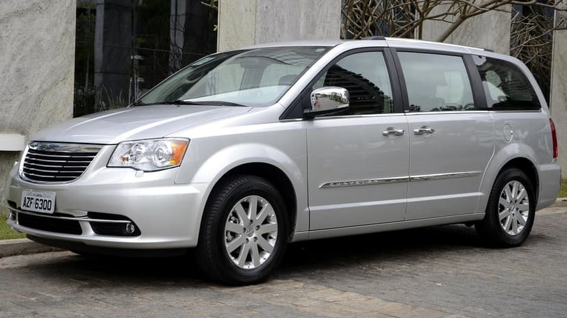 Silver Chrysler minivan parked on a stone driveway against a modern stone building in soft daylight.