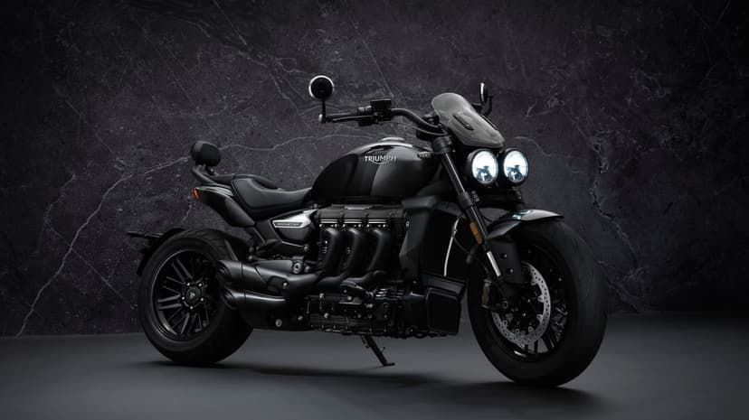 Matte black Triumph Rocket 3 GT motorcycle with glowing headlights against a dark textured stone wall.