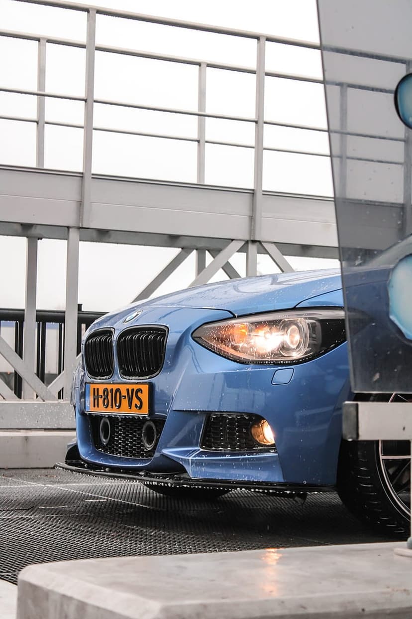 Blue BMW M Sport car with water droplets on the hood and grille parked at a metallic car wash station.