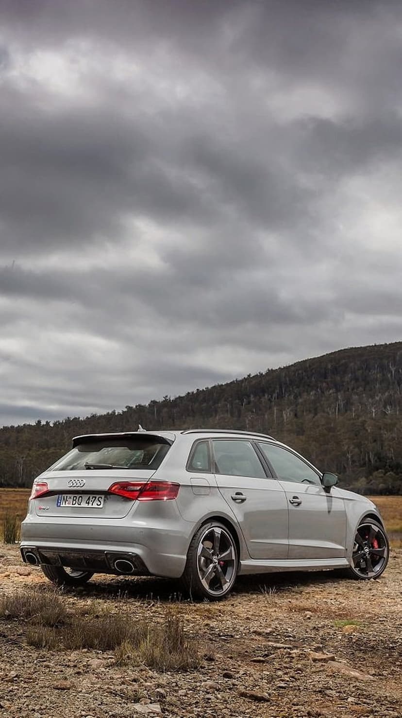 Audi RS3 Sportback in silver with black wheels parked on rocky terrain before a dense forest and cloudy sky.