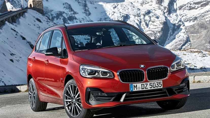 Metallic orange BMW parked on a winding alpine road with vast snow-covered mountain peaks in the background.