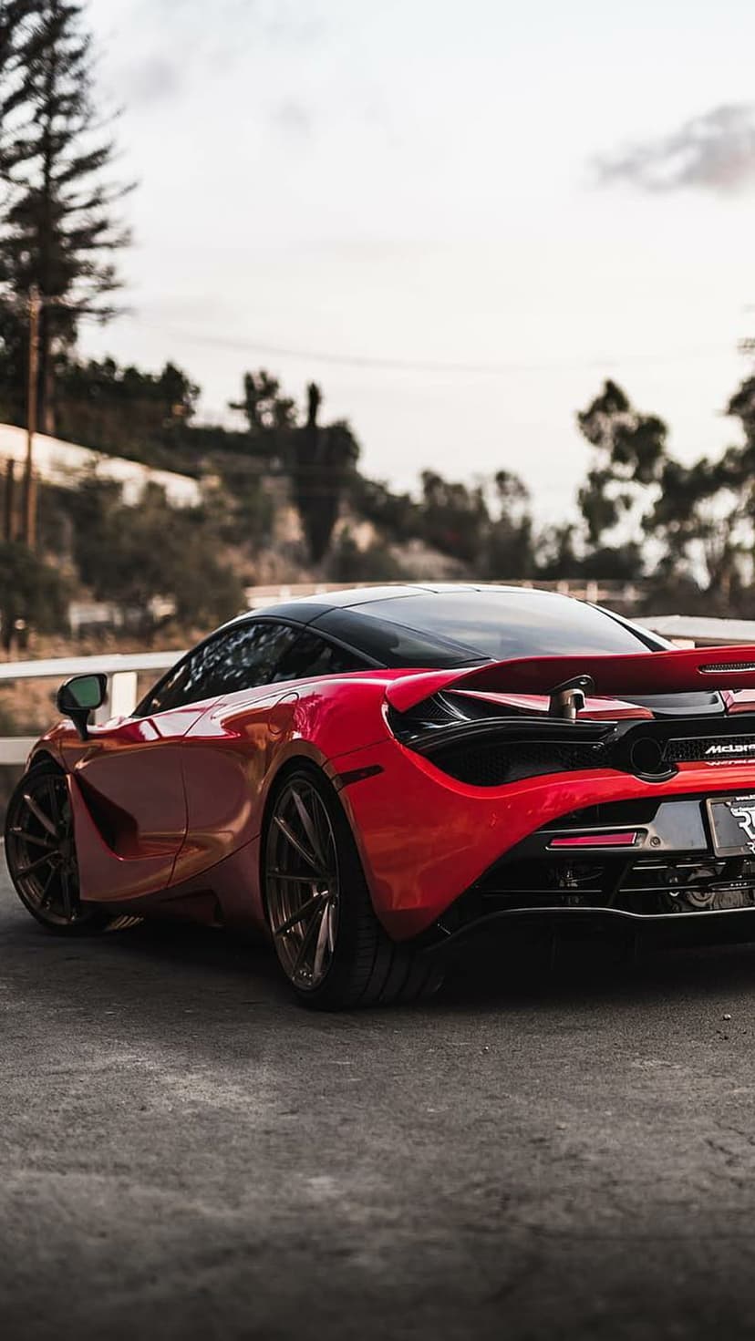 Red McLaren supercar with a rear spoiler and black roof sits against a blurred forest and overcast sky background.