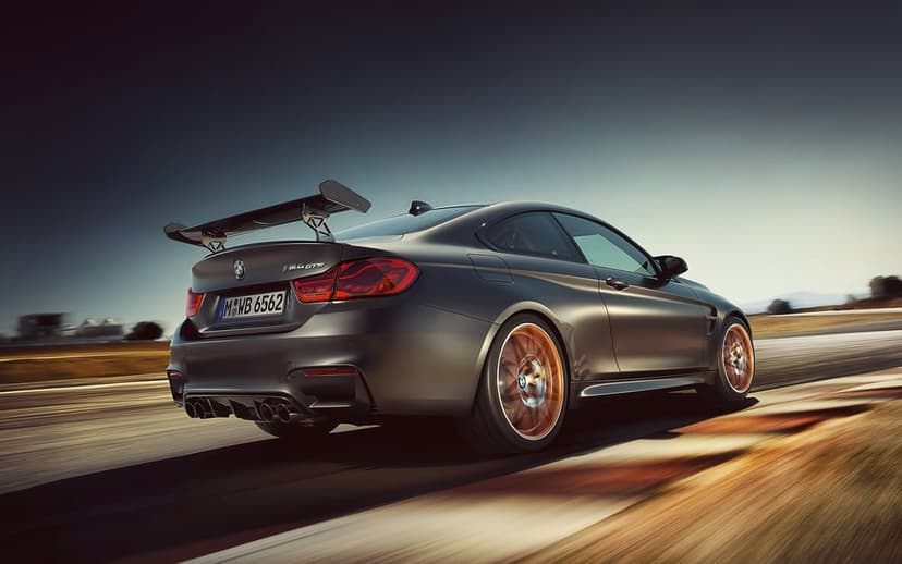 Matte grey BMW M4 GTS sports car with orange rims racing on a track with motion blur under a dramatic dark sky.