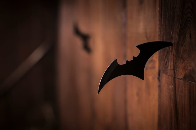 Black bat throwing star lodged in textured dark wood with a blurred shuriken in the shadowy background.