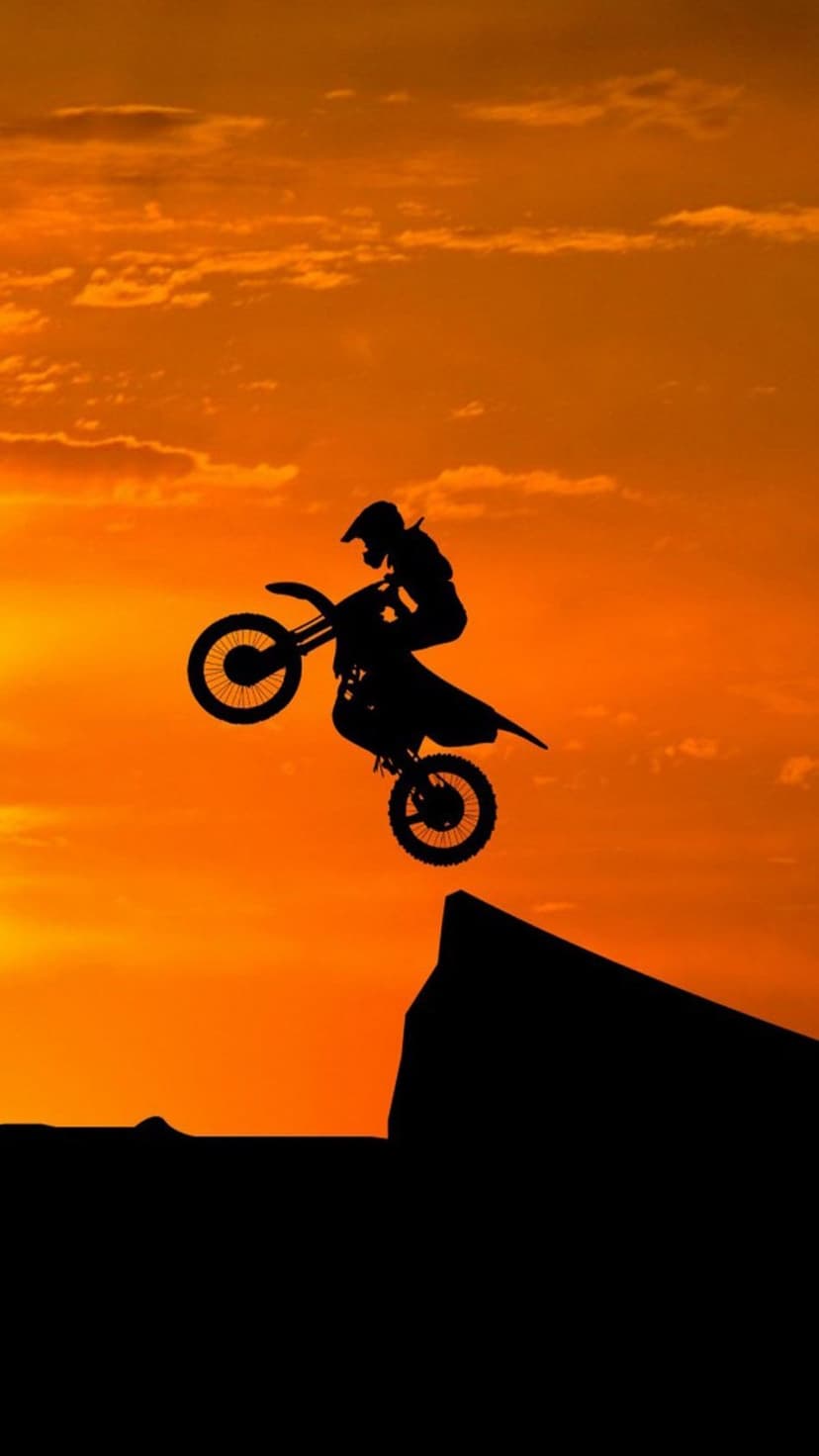 Motocross rider performs a high jump against a vivid orange sunset with wispy clouds in the background sky.