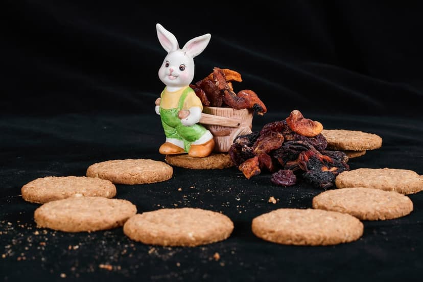 Ceramic bunny figurine standing by a wooden cart of dried apricots and prunes surrounded by golden oat biscuits.