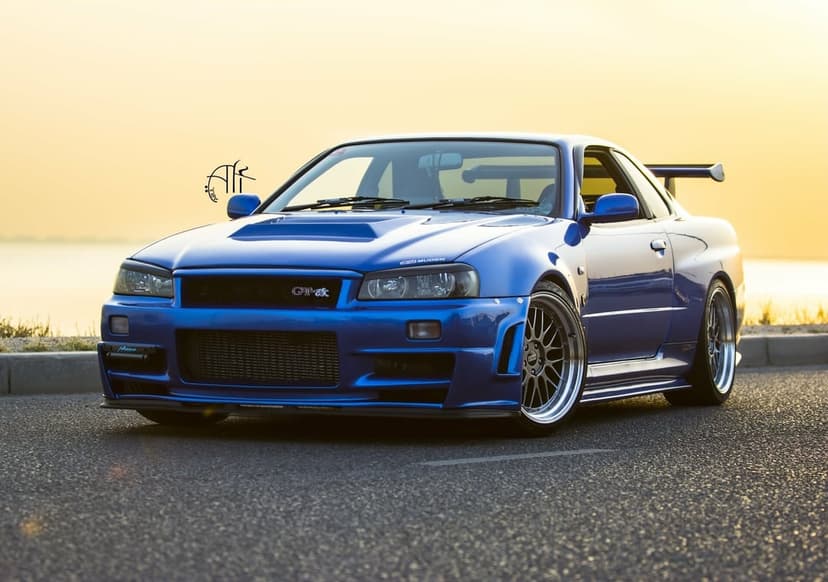 Blue Nissan Skyline GT-R R34 with custom wheels parked on an asphalt road during a warm golden hour sunset.