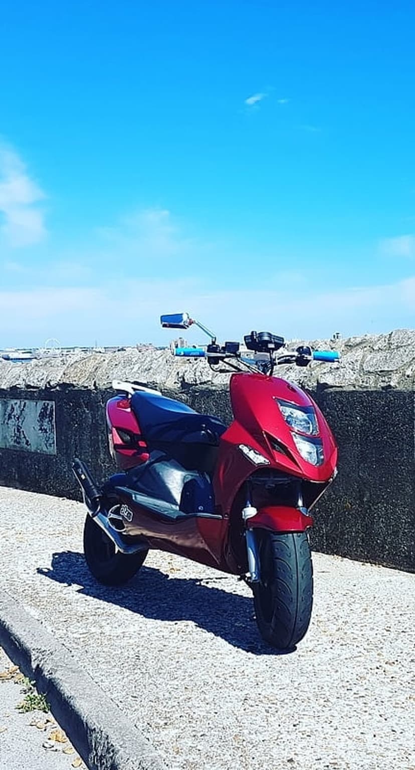 Red vintage scooter with chrome details parked by a grey stone wall under a bright blue sky with wispy clouds.