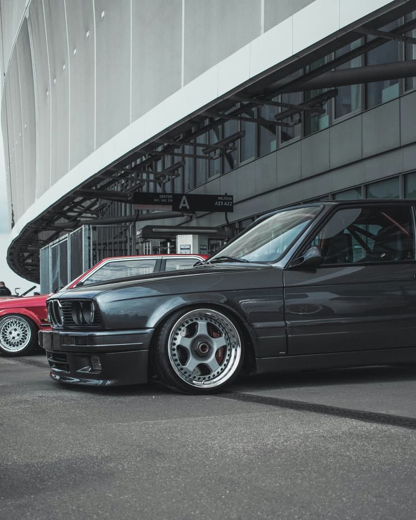 Dark gray BMW E30 M3 with chrome rims parked in front of a curved white modern building with glass windows.