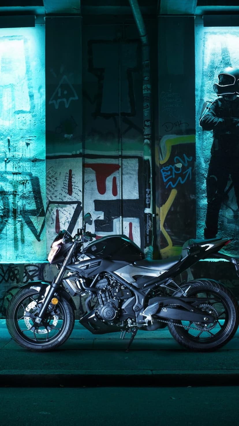 Black motorcycle parked in a dark urban alleyway illuminated by blue neon signs with graffiti on concrete walls.