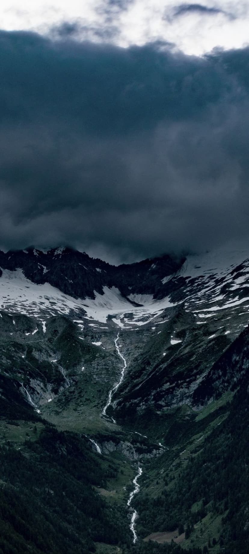 Snowy mountain peaks loom over a cascading waterfall under dark storm clouds within a lush green alpine valley.