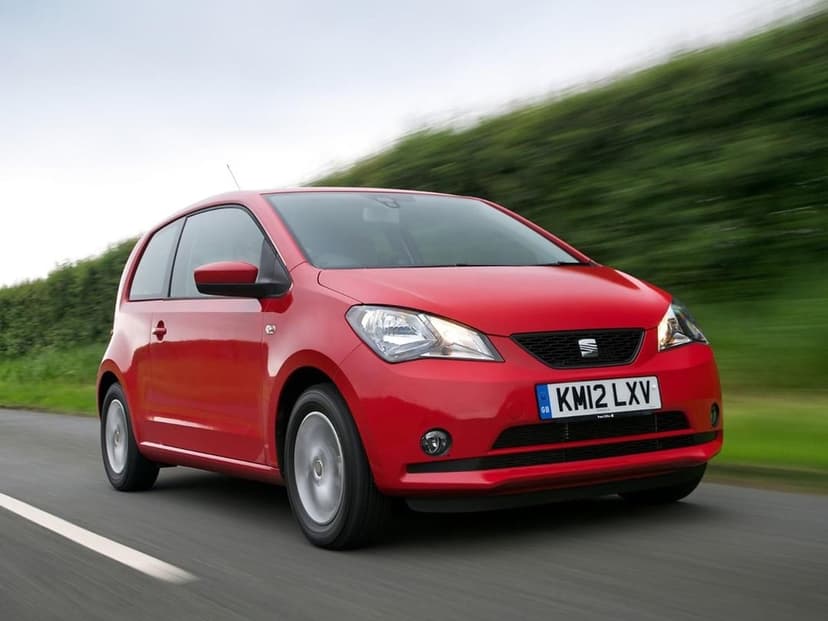 Red SEAT Mii hatchback driving on a road with motion blurred green hedges and a cloudy sky background.