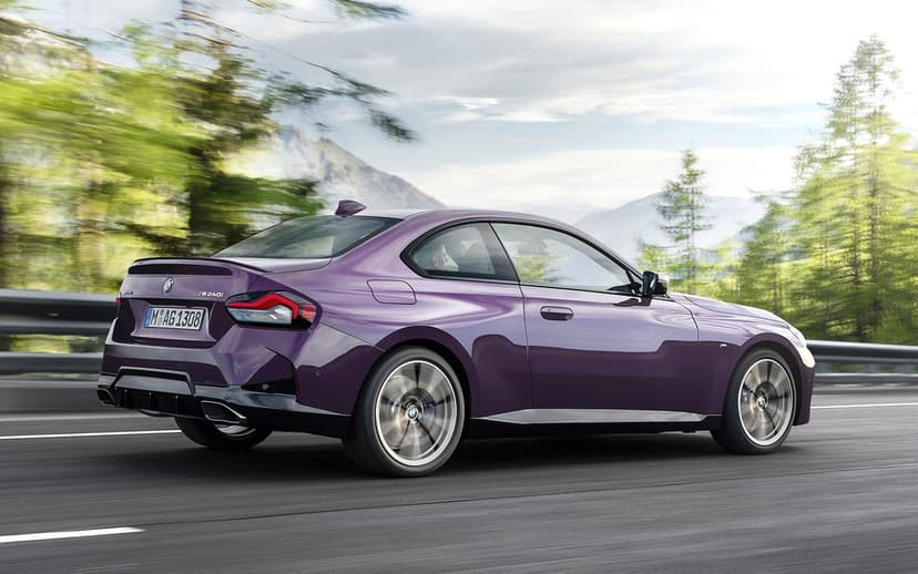 Purple BMW M240i sports coupe driving fast on a mountain road with motion blur and pine trees.
