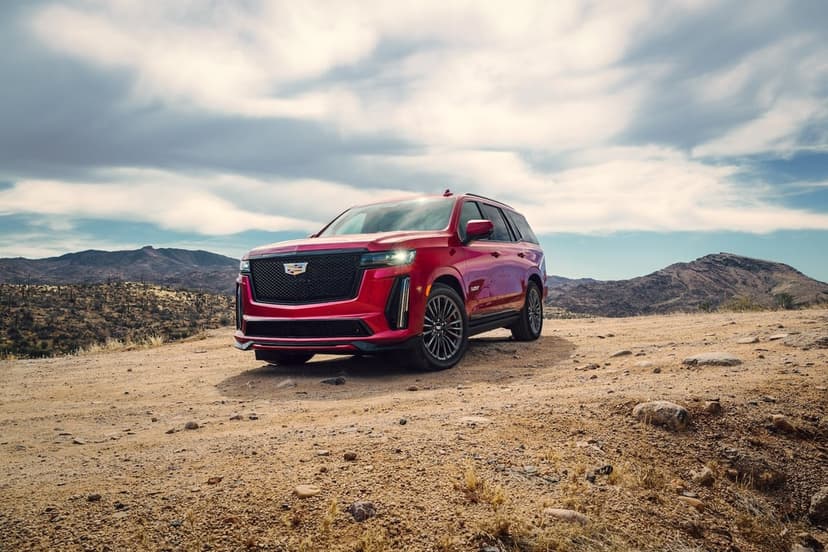 Red Cadillac Escalade-V SUV parked on a dusty desert trail under a vast cloudy sky with rocky hills behind.