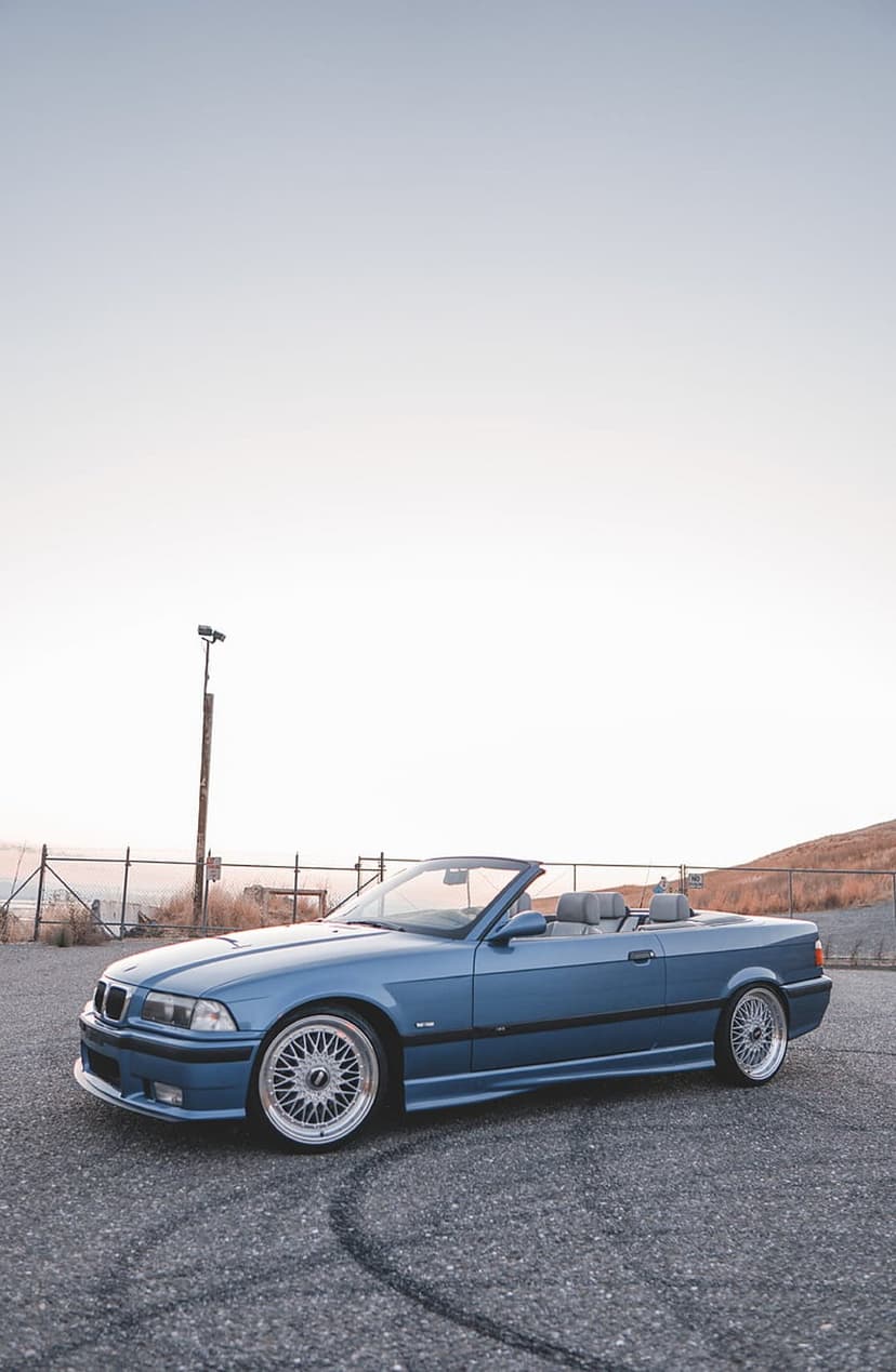 Blue BMW E36 convertible parked on rough asphalt under a pale sky during a hazy morning with visible tire marks.