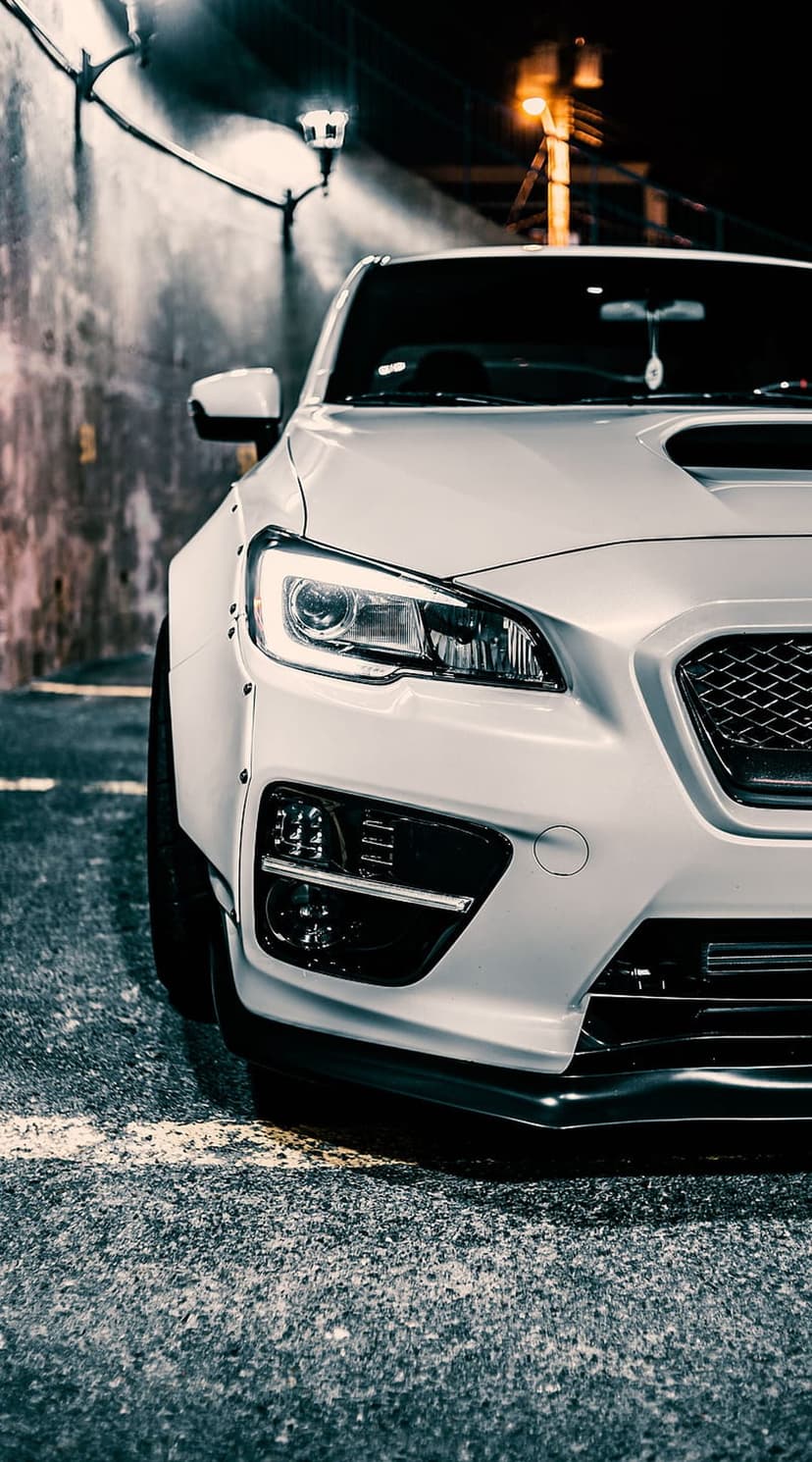 White Subaru WRX STI with glowing headlights and a widebody kit parked on a dark asphalt street at night.