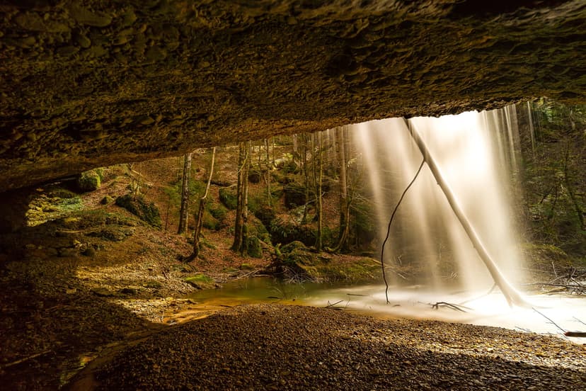 Cascading water drops from a mossy stone overhang into a calm pool inside a lush green forest cave.
