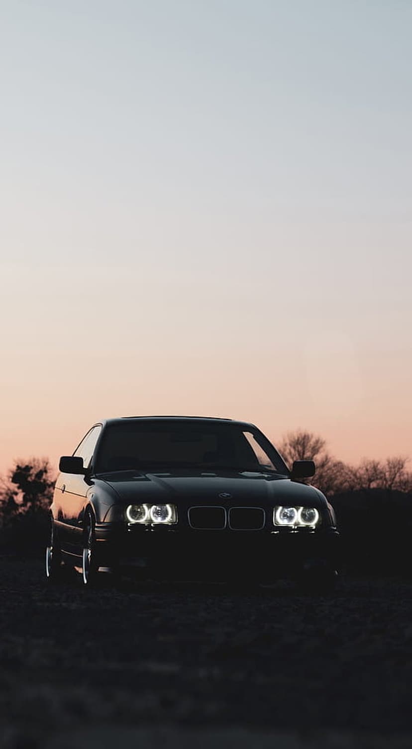 Black BMW E36 coupe with glowing halo headlights parked on rough terrain against a colorful gradient sunset sky.