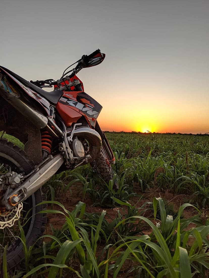 Dirt bike parked in a grassy field under a glowing orange sunset sky with long shadows across the landscape.