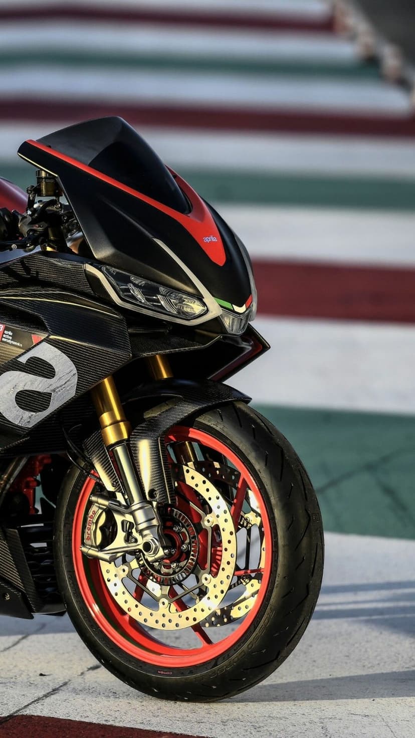 Close-up of an Aprilia motorcycle with carbon fiber bodywork, gold forks, and red wheels on a race track.