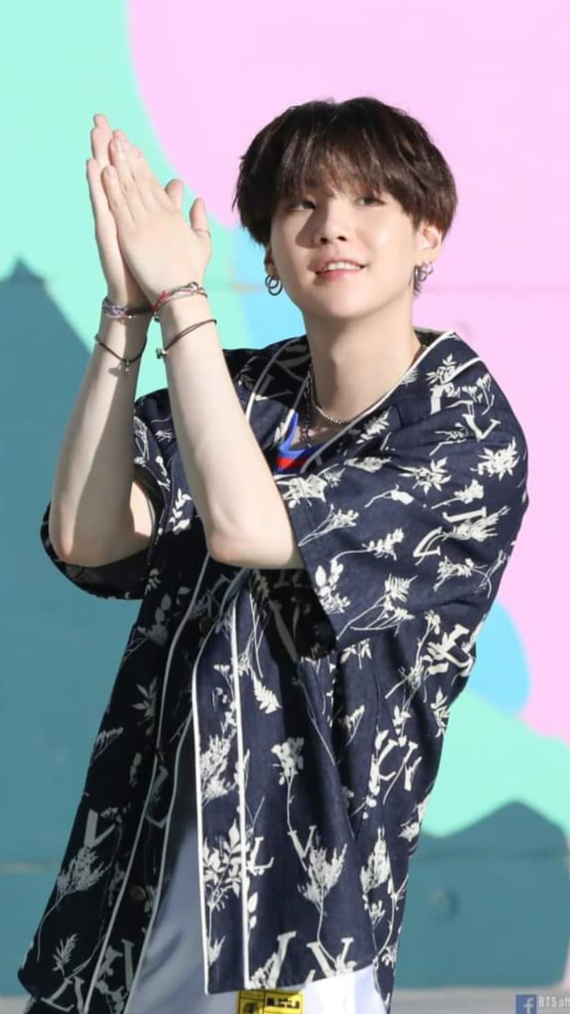 Suga of BTS clapping while wearing a navy floral print shirt against a teal and pink gradient backdrop.