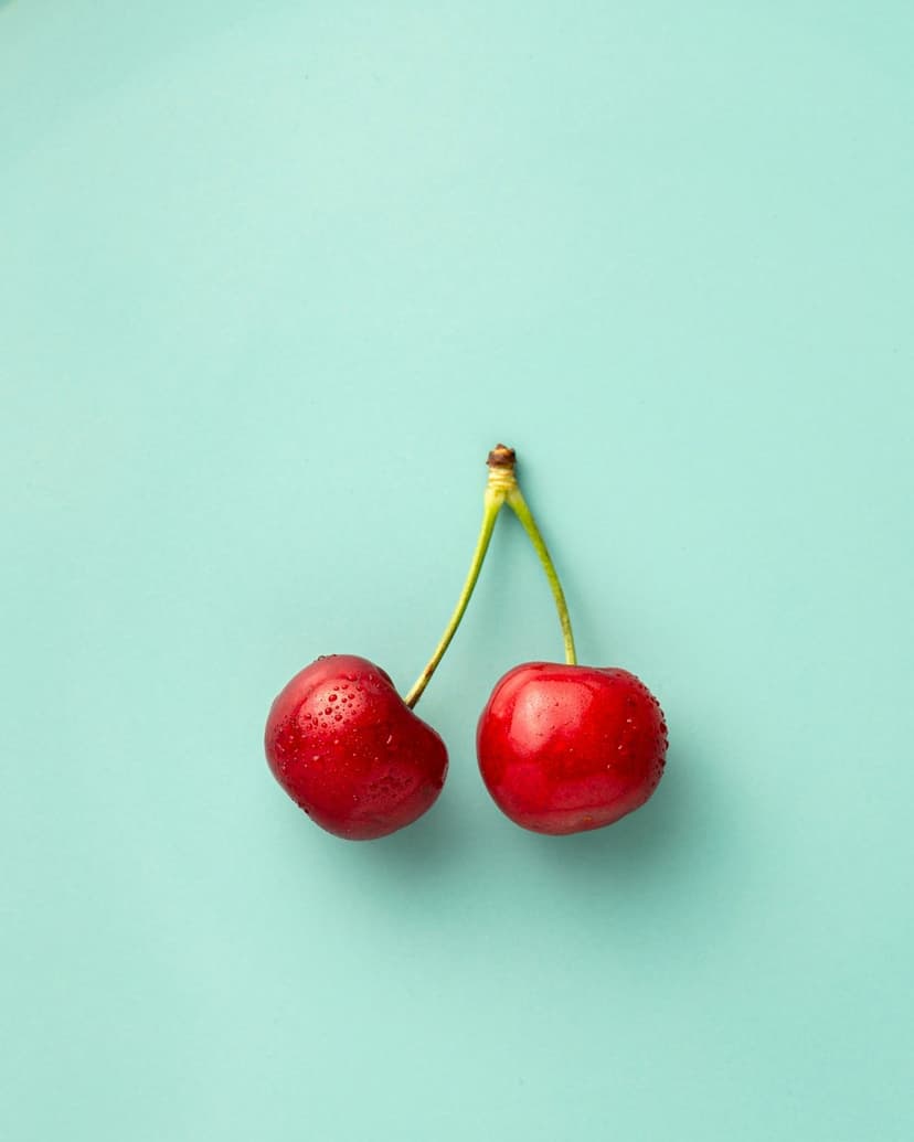 Two red cherries with glistening water droplets sit on a smooth light blue surface in a minimalist macro view.