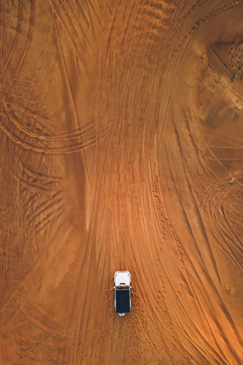 Monochrome vehicle driving through vast sand dunes leaving tire tracks across the golden desert surface.