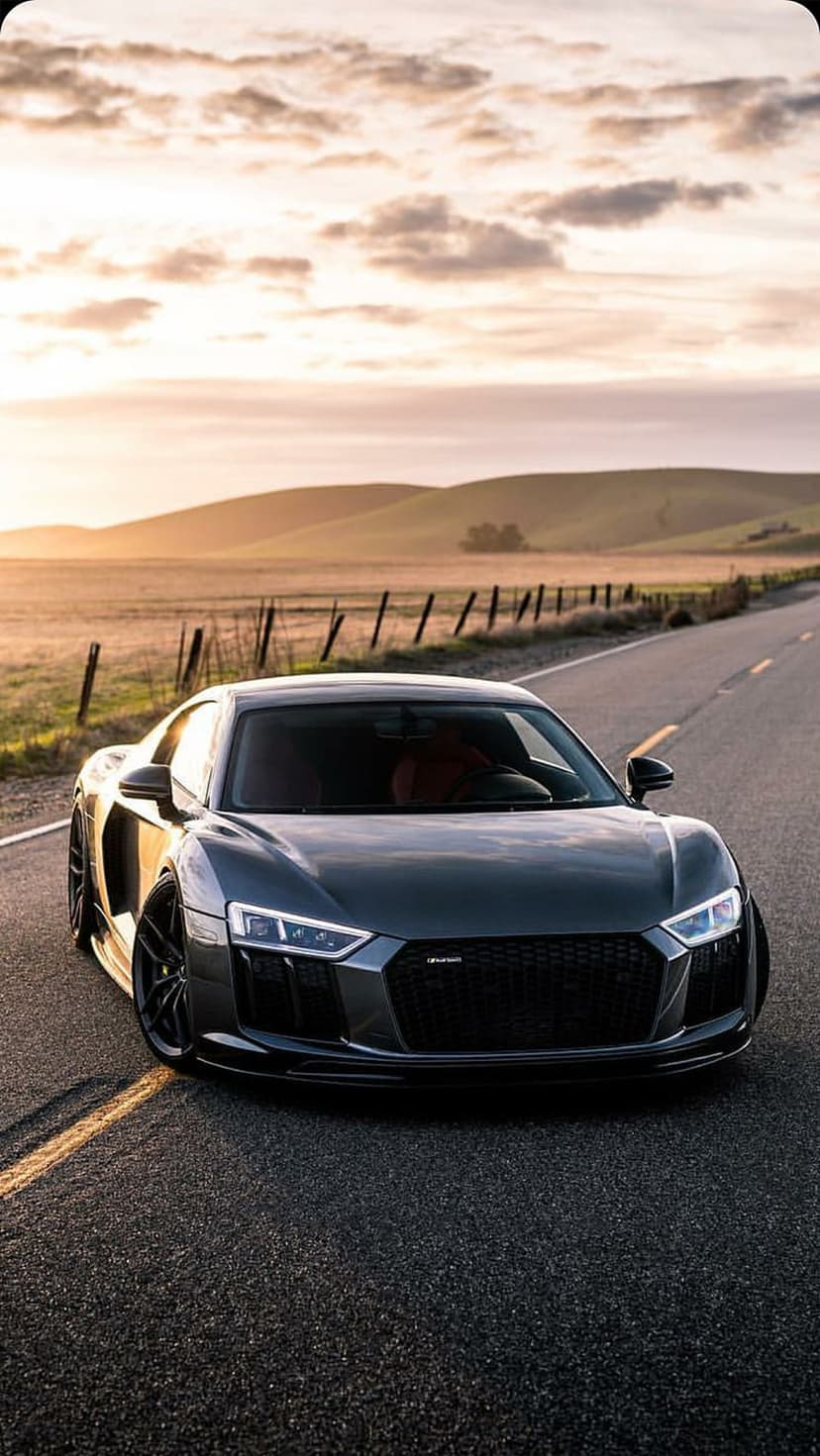 Audi R8 sports car parked on a winding road with rolling hills under a vibrant orange and purple sunset sky.