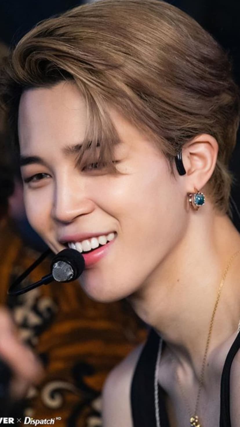Jimin from BTS smiling into a microphone while wearing a silver earring and gold chain necklace.
