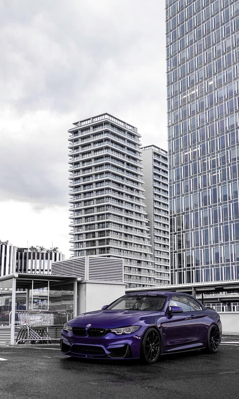 Purple BMW M4 coupe parked on wet asphalt before modern glass skyscrapers under a cloudy grey sky.