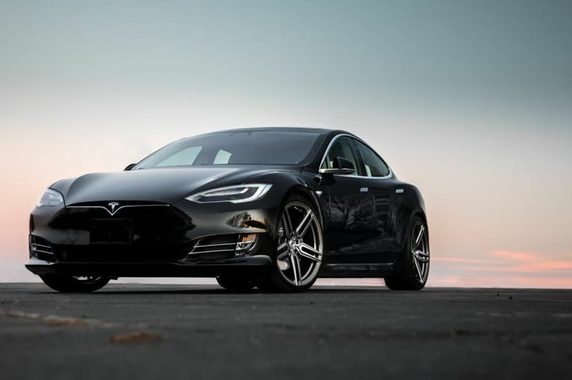 Black Tesla Model S luxury electric sedan parked on asphalt with custom rims against a soft sunset sky.
