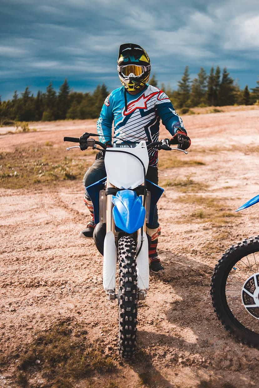 Motocross rider in full gear sits on a blue and white dirt bike on a dusty track under a dramatic cloudy sky.