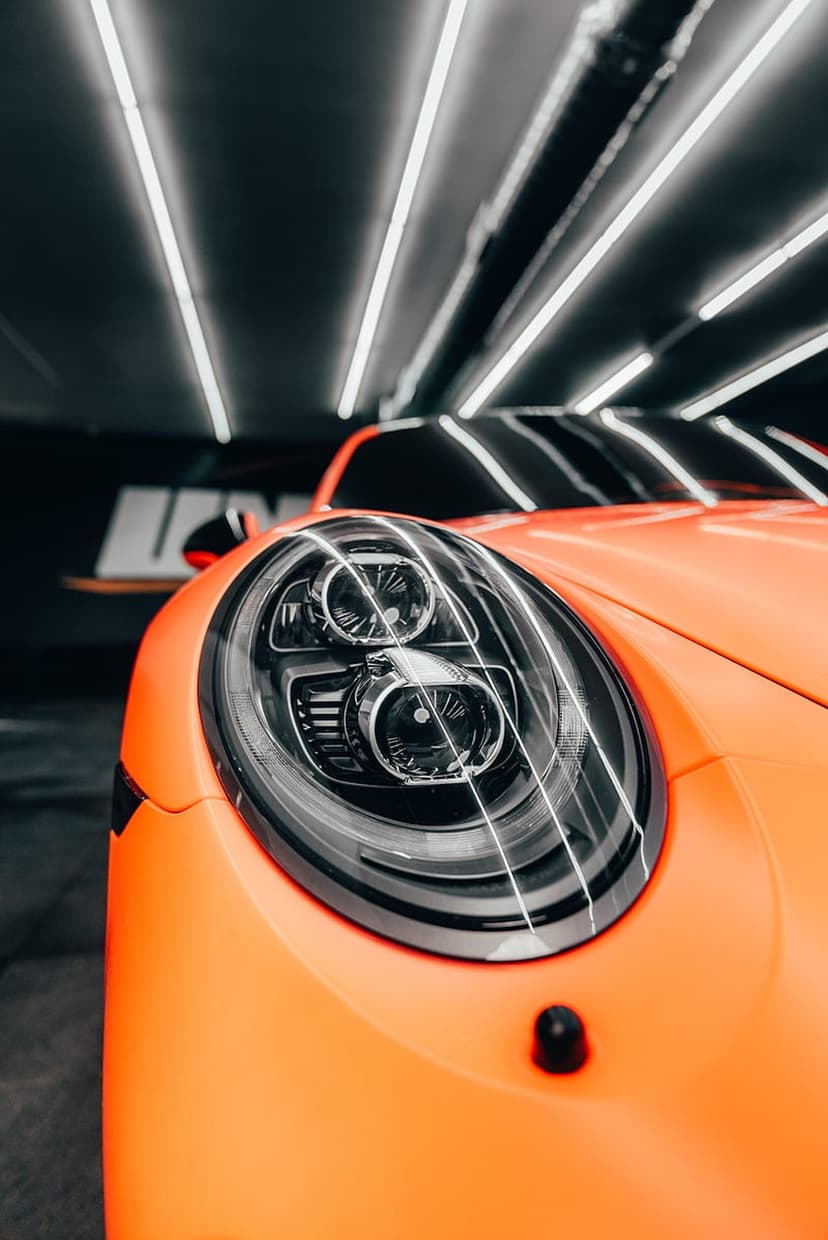 Porsche headlight assembly shows intricate glass optics and orange paint under linear overhead studio lights.